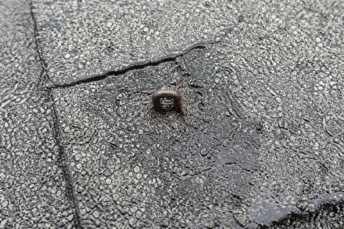 A rusted staple driven through the face of a weathered asphalt shingle, granules worn away and a dark moisture stain spreading from the puncture